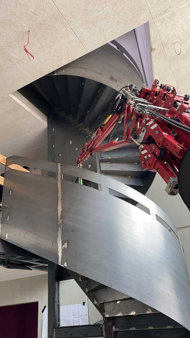 Hydraulischer Kran hebt Bauteile innerhalb eines engen Treppenhausschachtes.